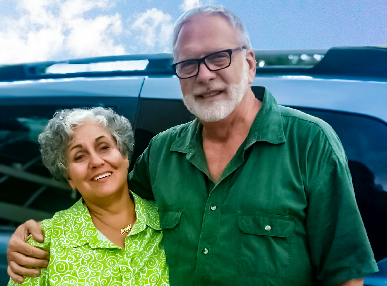 Melanie & Kirk, Owners of DummyEggs.com An older couple smiles warmly, the woman with curly gray hair and green blouse leaning on a man with glasses, white beard, and green shirt, standing before a car under a cloudy sky.